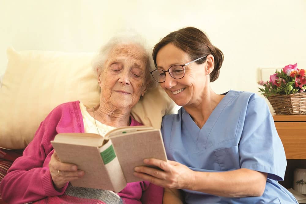 Right at Home Caregiver Seated on Bed Reading Book with Elderly Woman female carer reading a book with a senior female