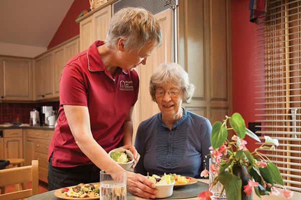 a female carer serving a meal to a senior female at the dining room table