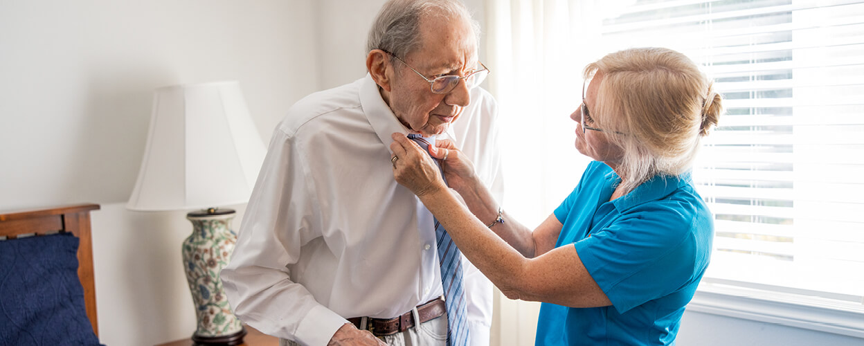 Female caregiver helping senior male with his tie in a bedroom near a window