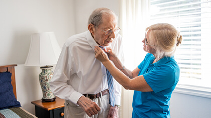 Female caregiver helping senior male with his tie in a bedroom near a window