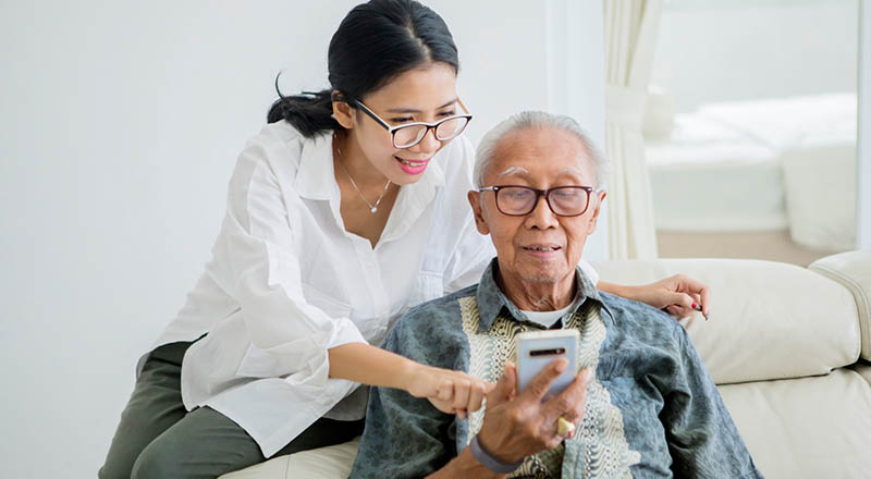 A senior male is wearing a pacsana bracelet and looking at a cell phone while his female daughter is looking over his shoulder